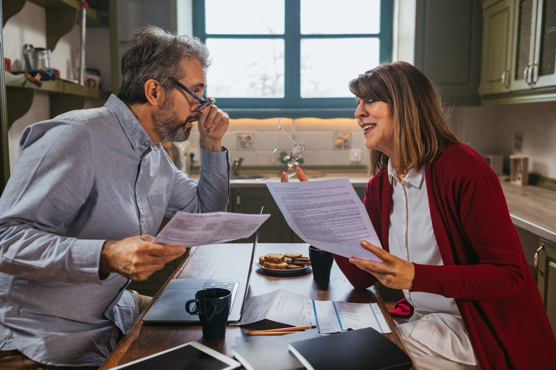 Couple reviewing paperwork at a kitchen table. Woman smiles, holding papers, man looks at papers and glasses.