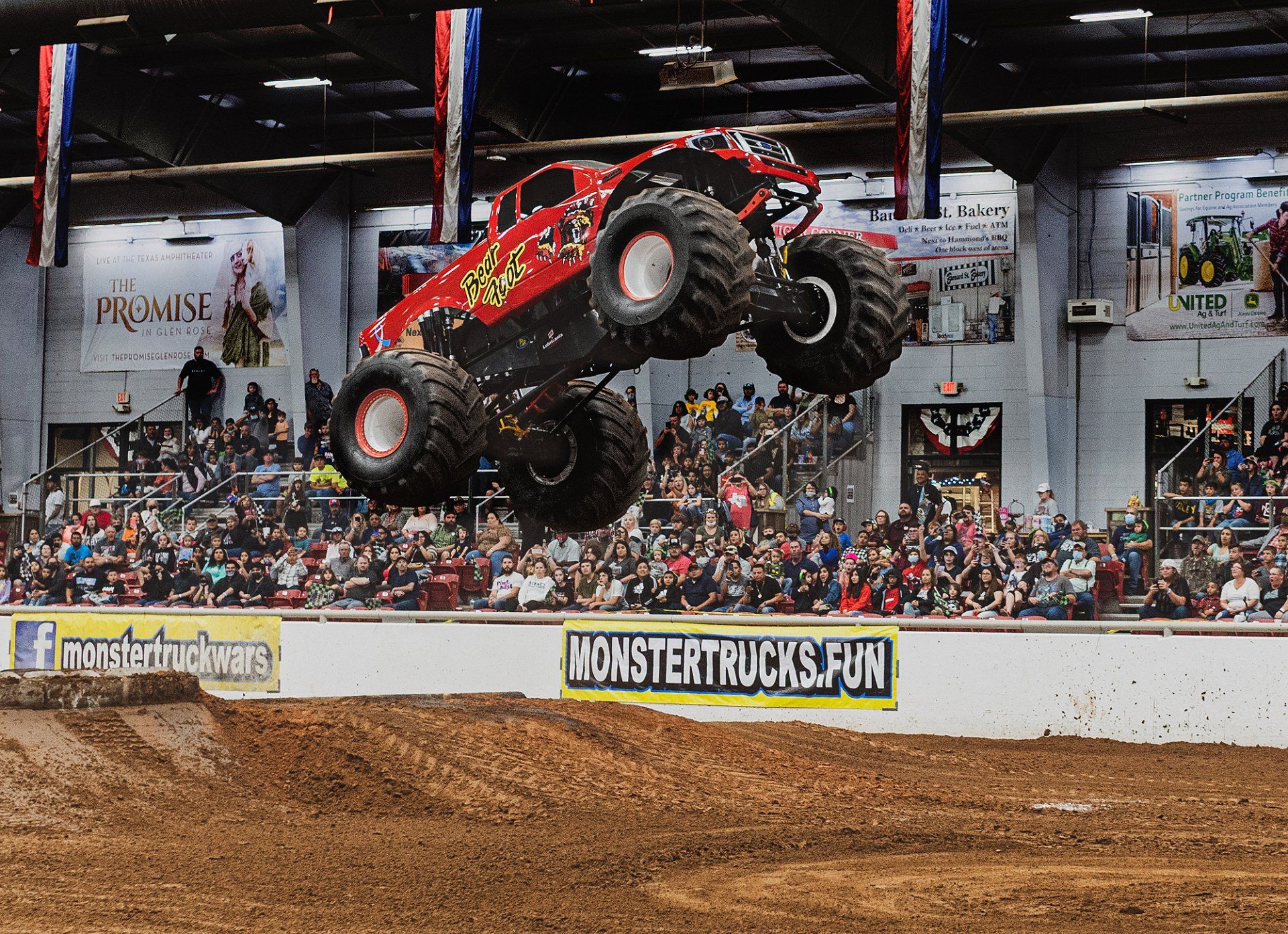 Monster truck in mid-air, jumping over a dirt mound in an arena filled with spectators.