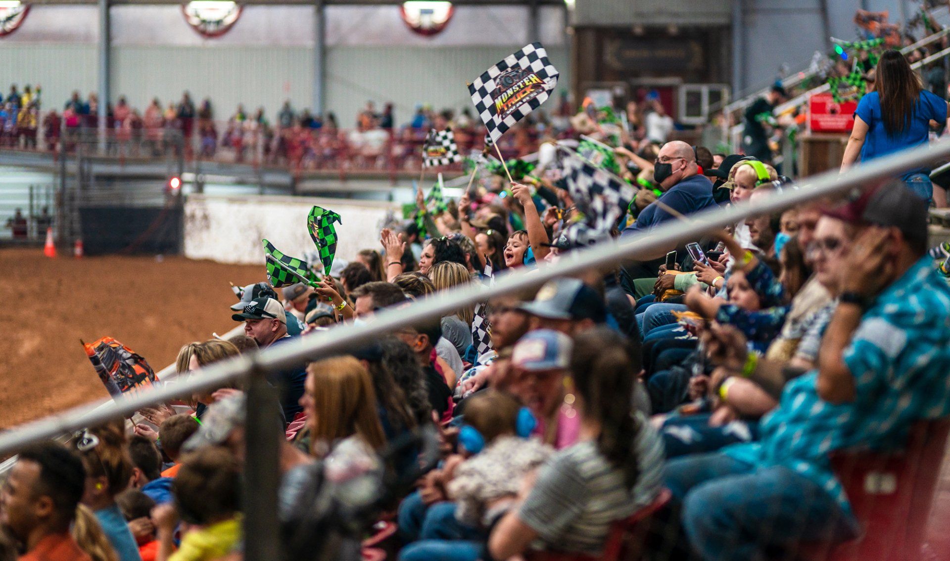 Crowd watches a race, waving checkered flags. Indoor arena with dirt track.