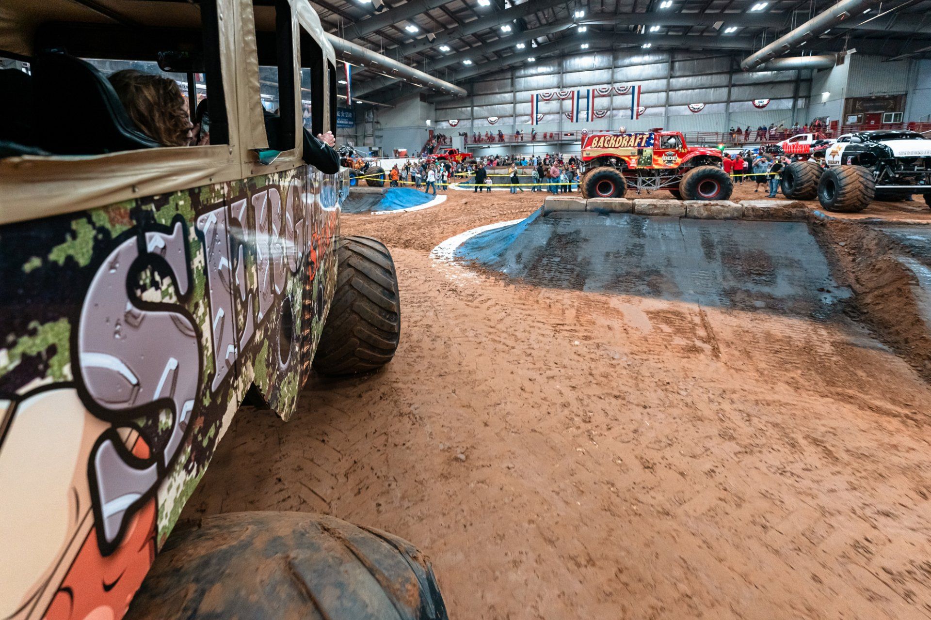 Monster truck event: trucks on dirt course, spectators in background.
