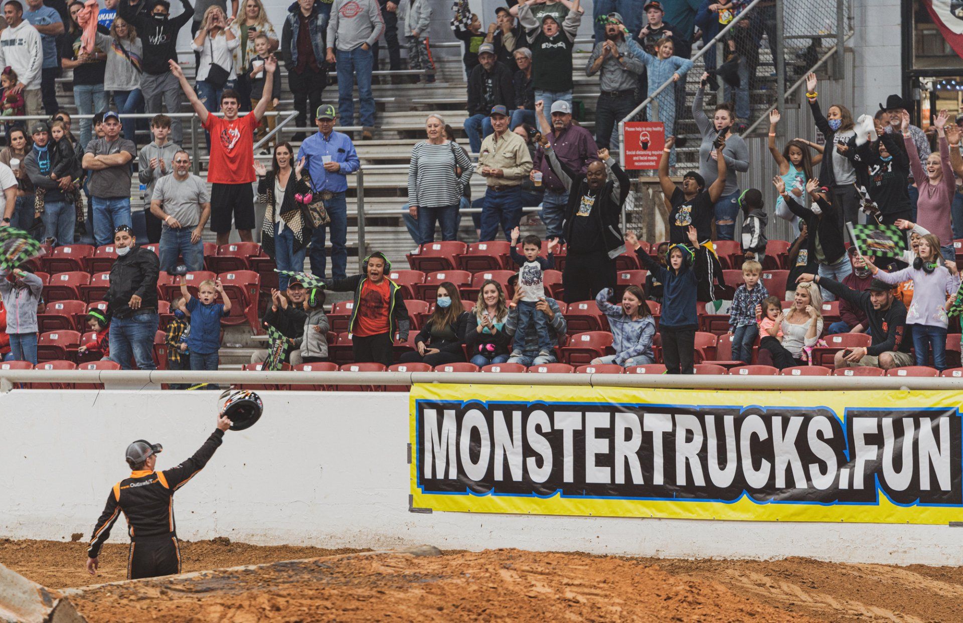 Monster truck driver waves to cheering crowd at an arena, a banner reads