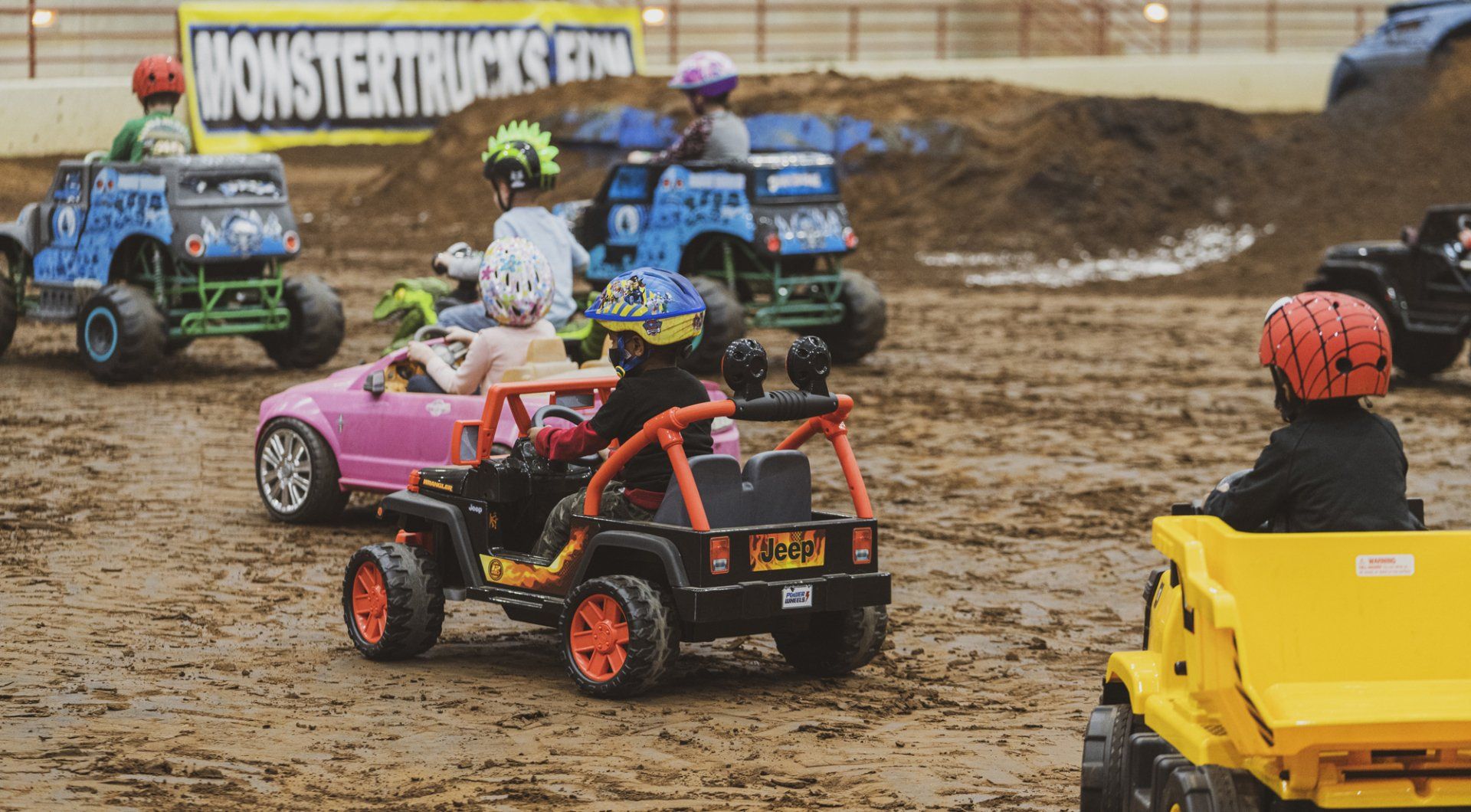 Children in toy trucks on a muddy track, racing. Yellow, blue, pink, and black vehicles; helmets worn.