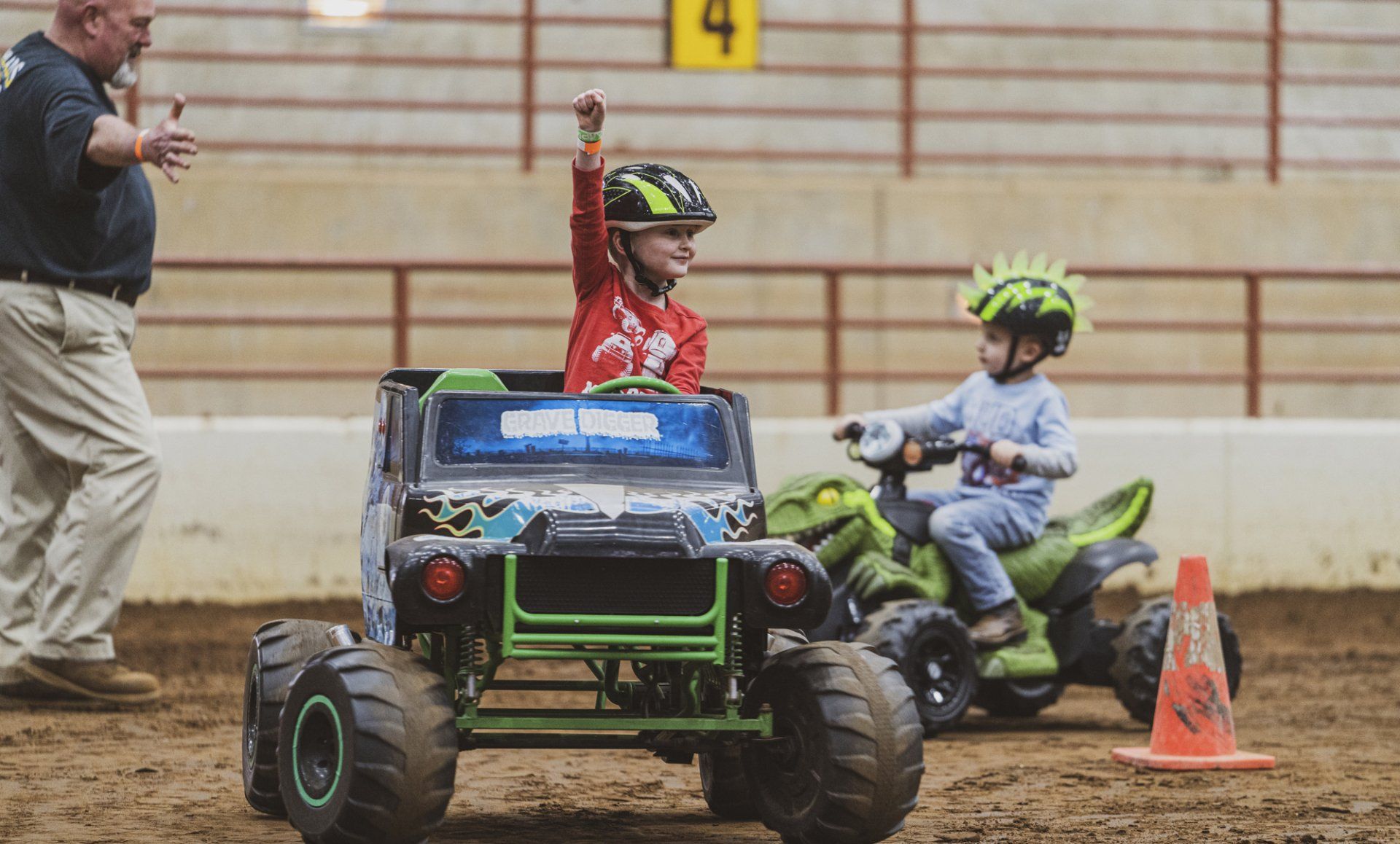 Two children on toy vehicles compete in a race; one raises a hand in victory. Indoors, dirt track, adult watches.