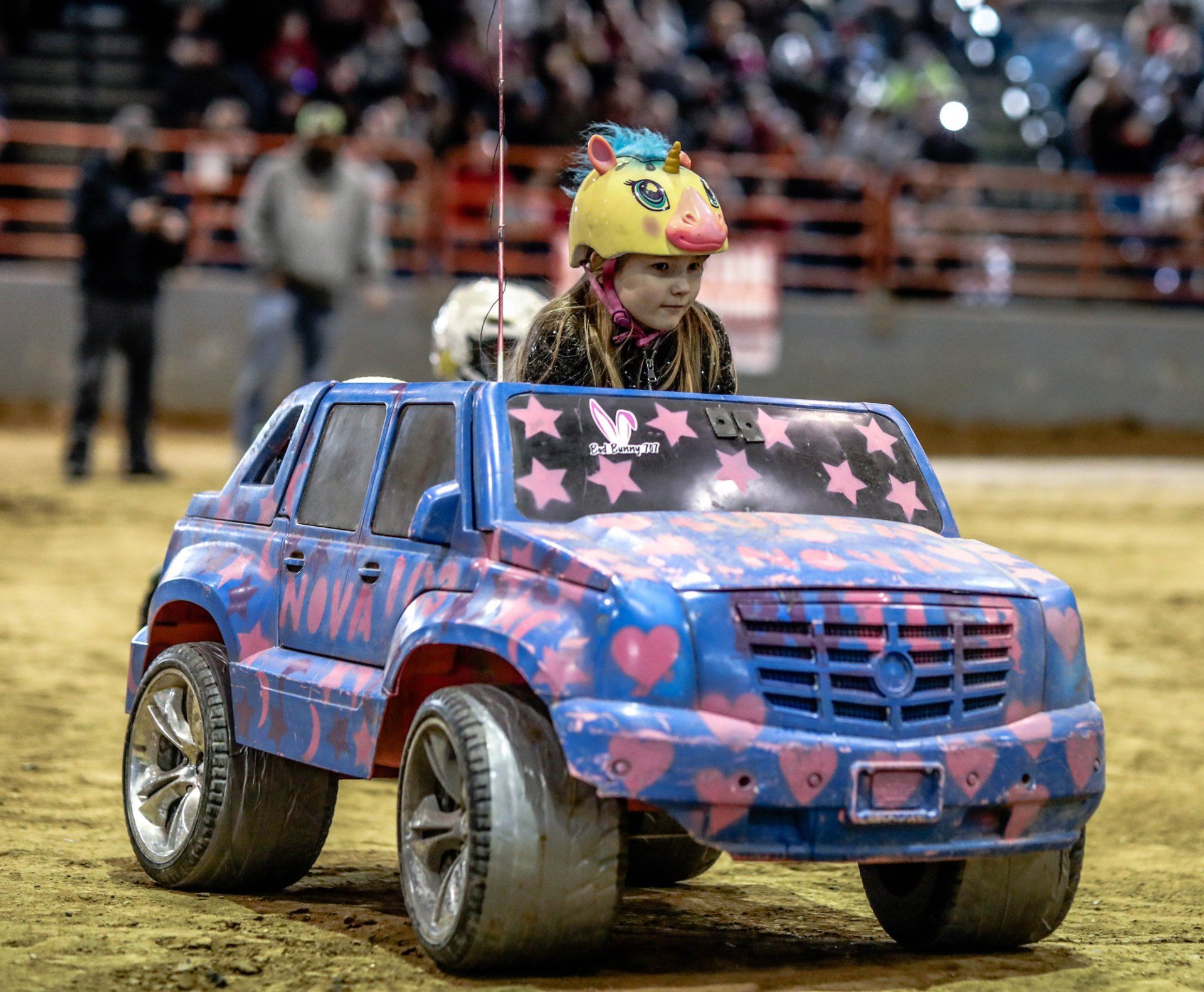 Child in a decorated toy car, wearing a unicorn helmet, racing on a dirt track.