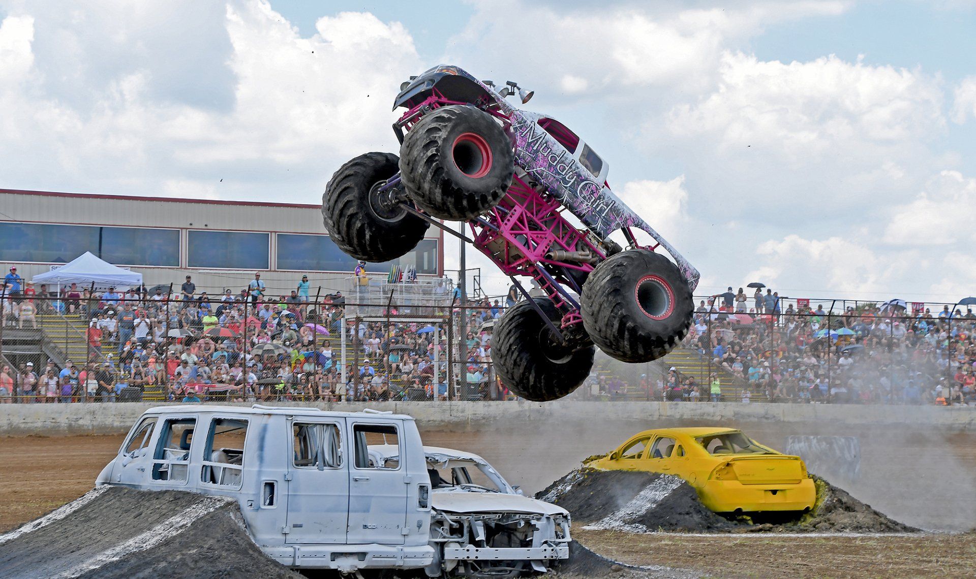 Monster truck, pink and black, airborne over crushed cars at a stadium event.