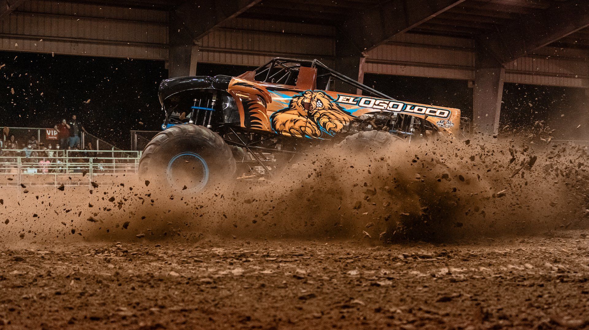 Monster truck, kicking up dirt, performing in arena. Black and orange paint scheme.