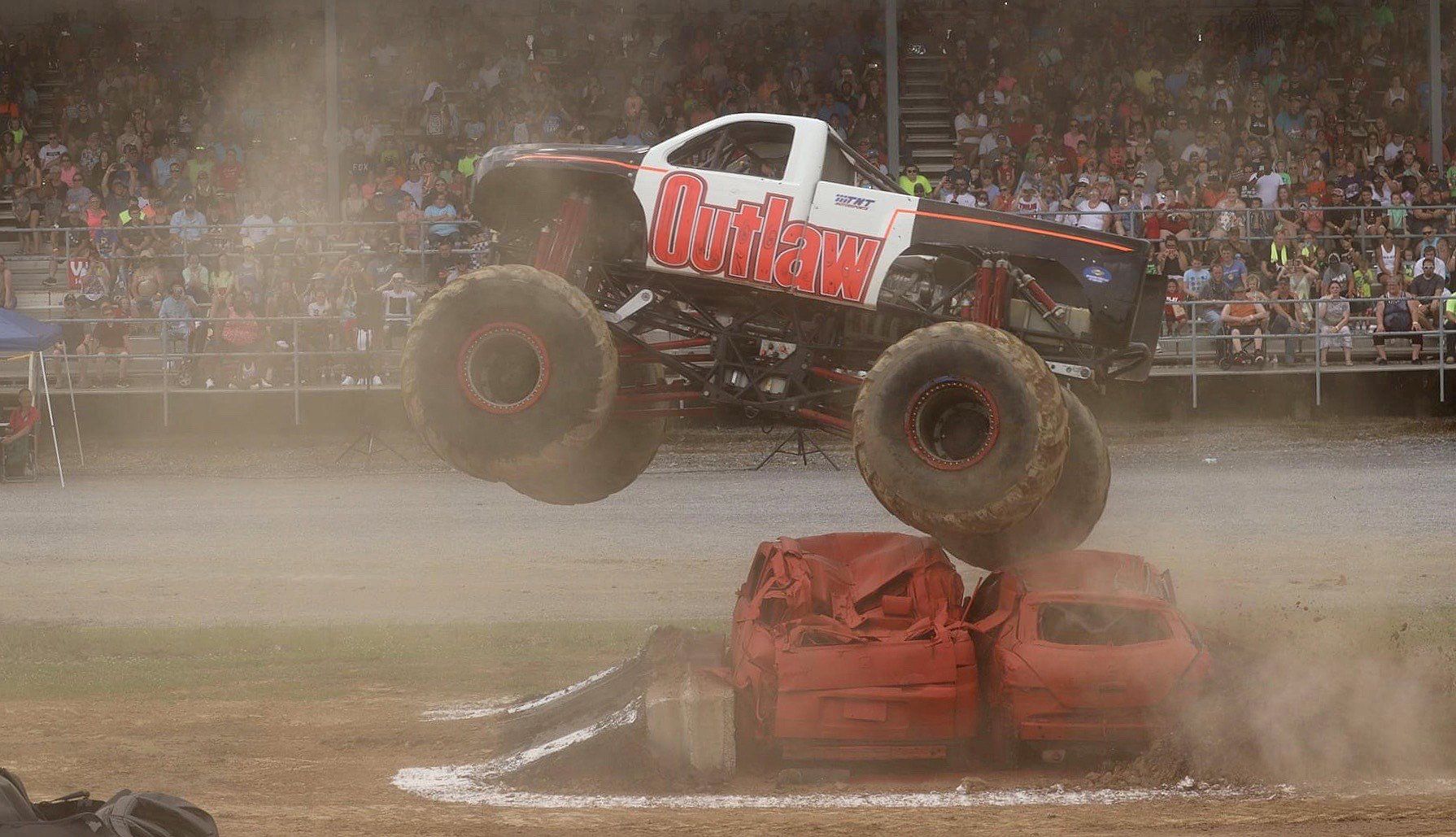 Monster truck, red and white, jumps over two crushed cars at an arena show.