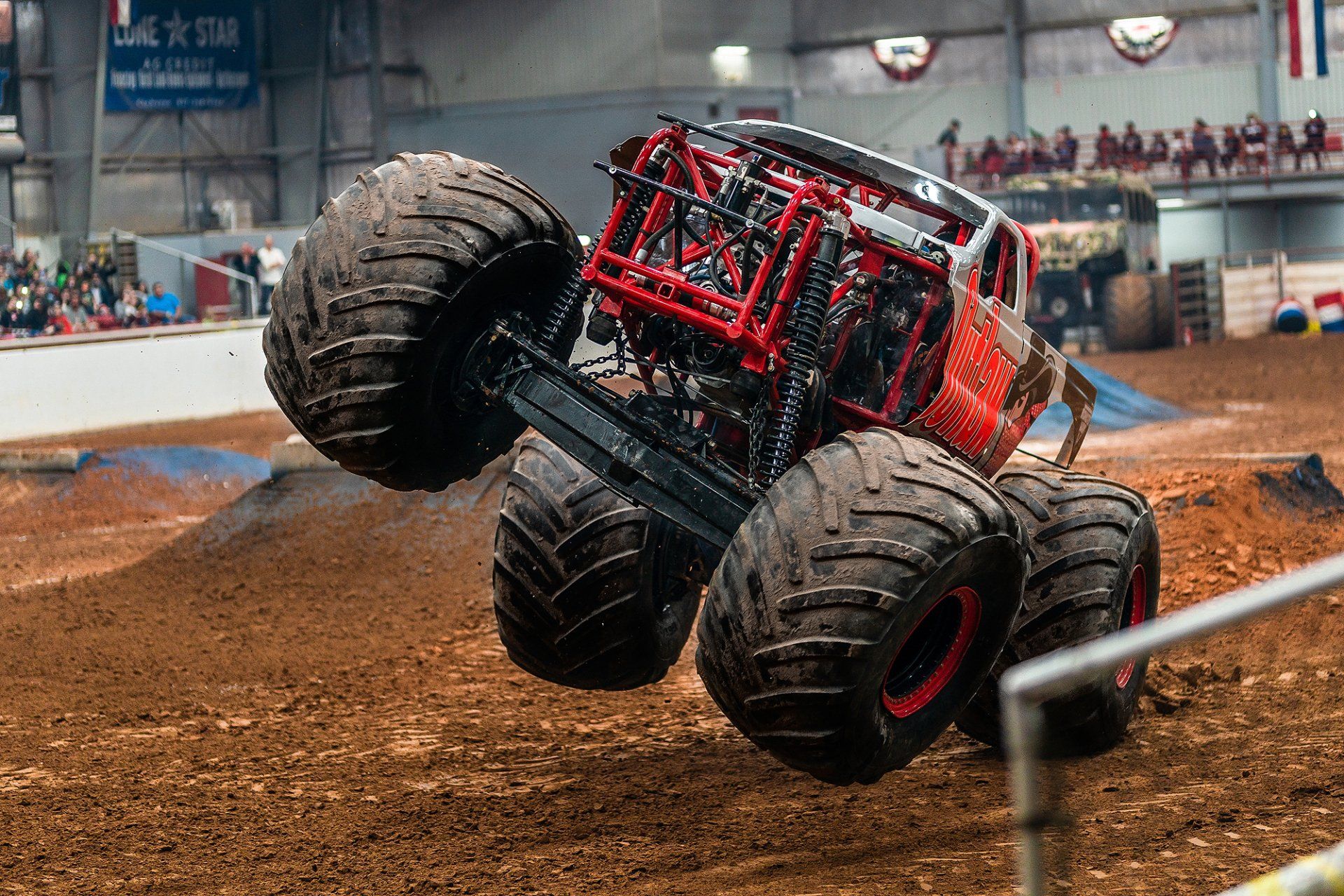 Monster truck, red and white, in mid-air over a dirt track. Black tires, spectators in the background.