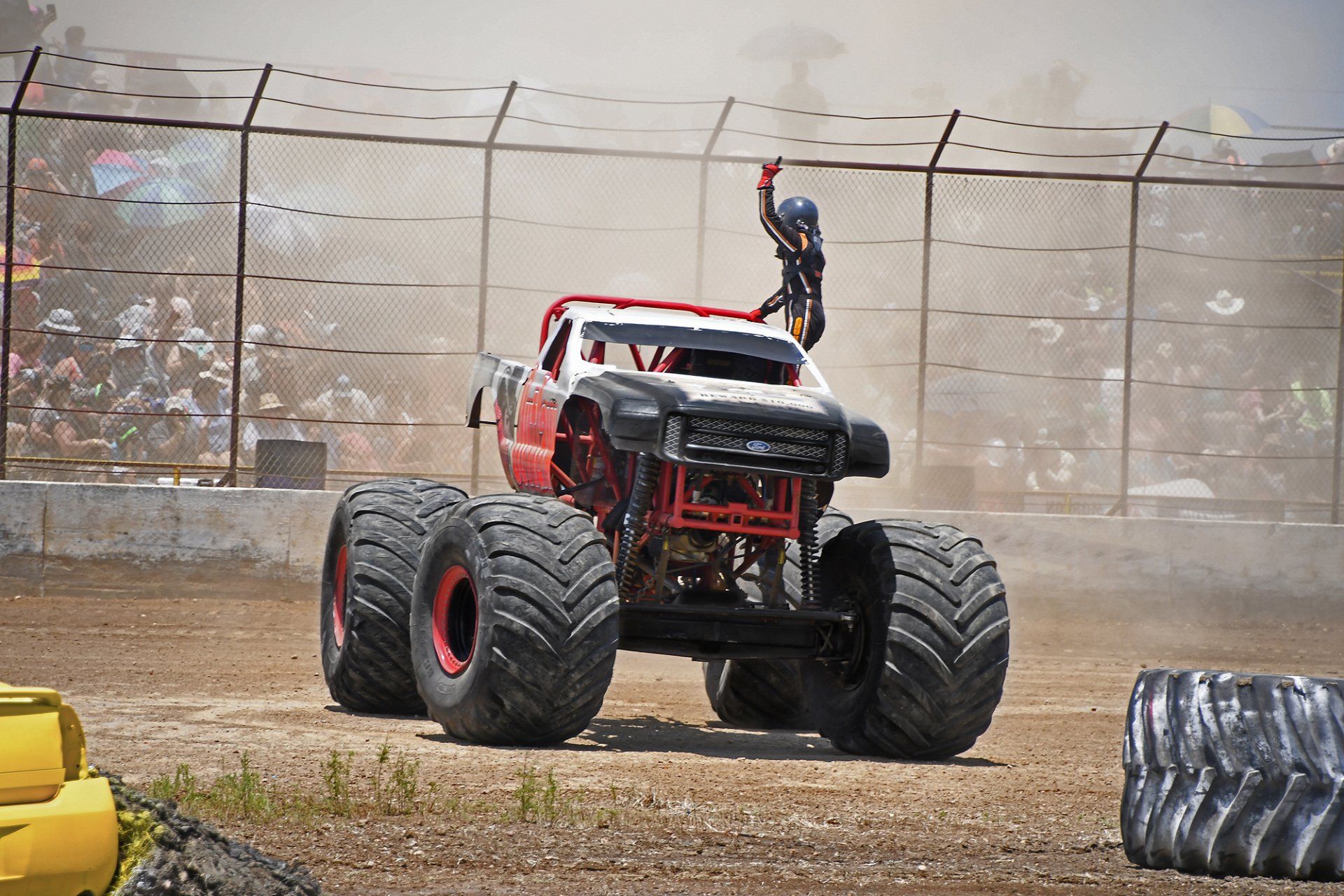 Monster truck in a dirt arena, driver waving from the roof, cheering crowd in the background.