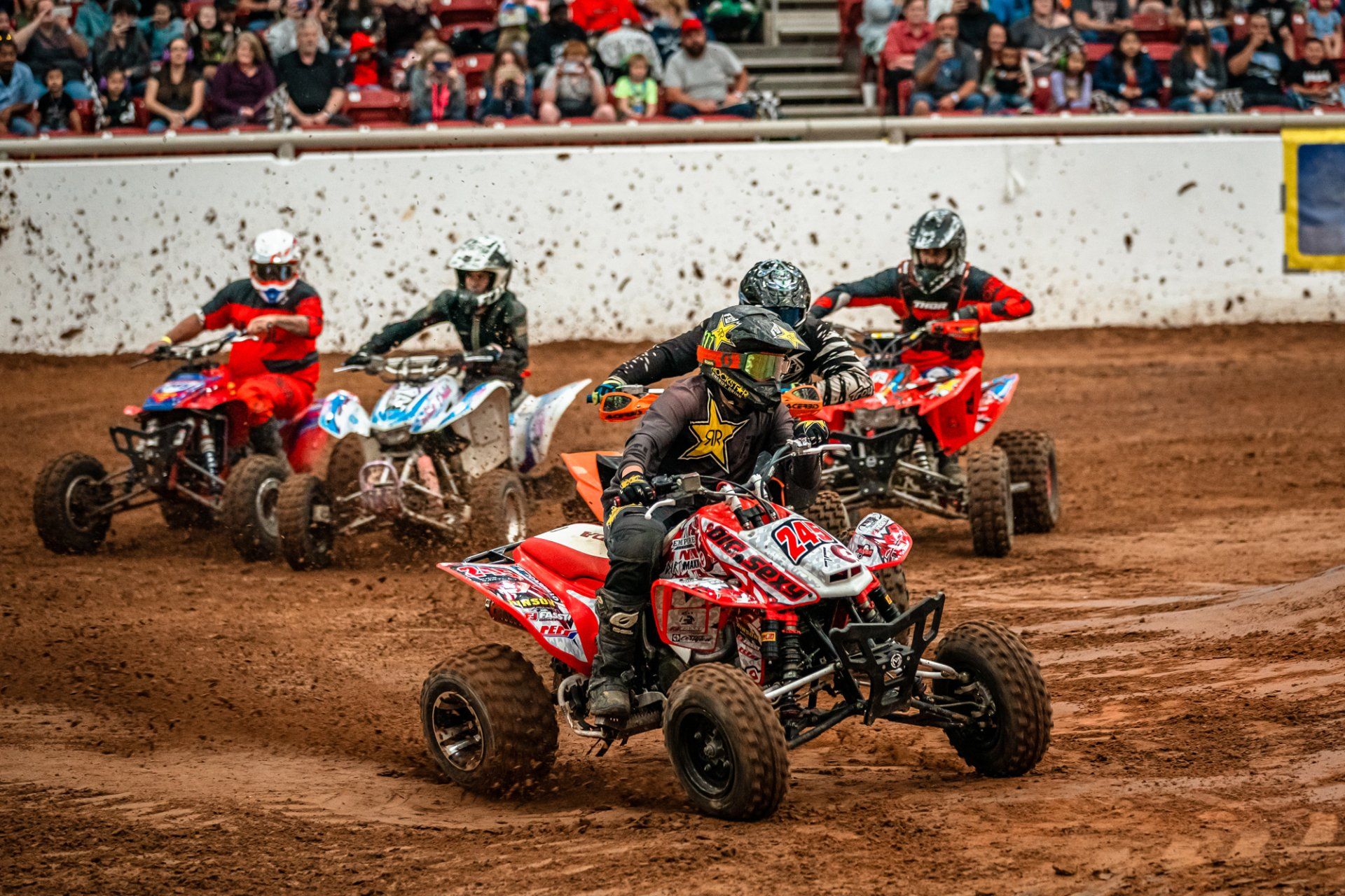 ATV racers speeding around a dirt track, spectators in the background.