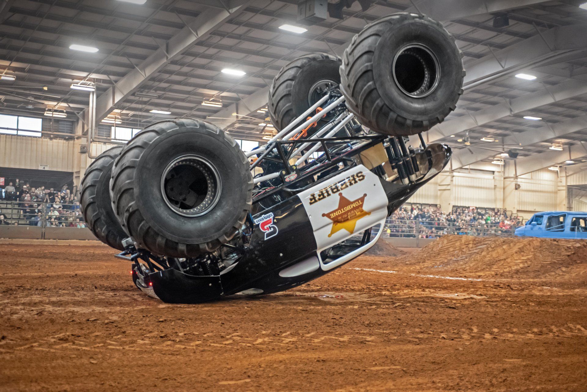 Monster truck Sheriff performing a stunt, on its side. Arena setting with spectators.