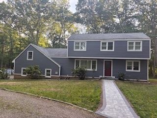Gray two-story house with white trim, red front door, and a stone walkway in a wooded yard.