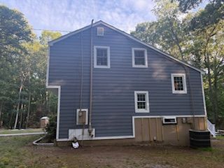 Blue two-story house with white trim, small basement windows, and trees in the background