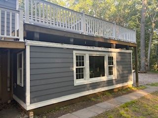 Gray house exterior with white-trimmed windows and upper deck, beside a wooded yard
