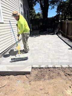 Worker leveling light gray pavers in a narrow backyard patio beside a house