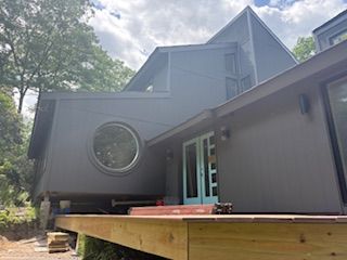 Modern dark gray house with a circular window and a raised wooden deck in a wooded setting