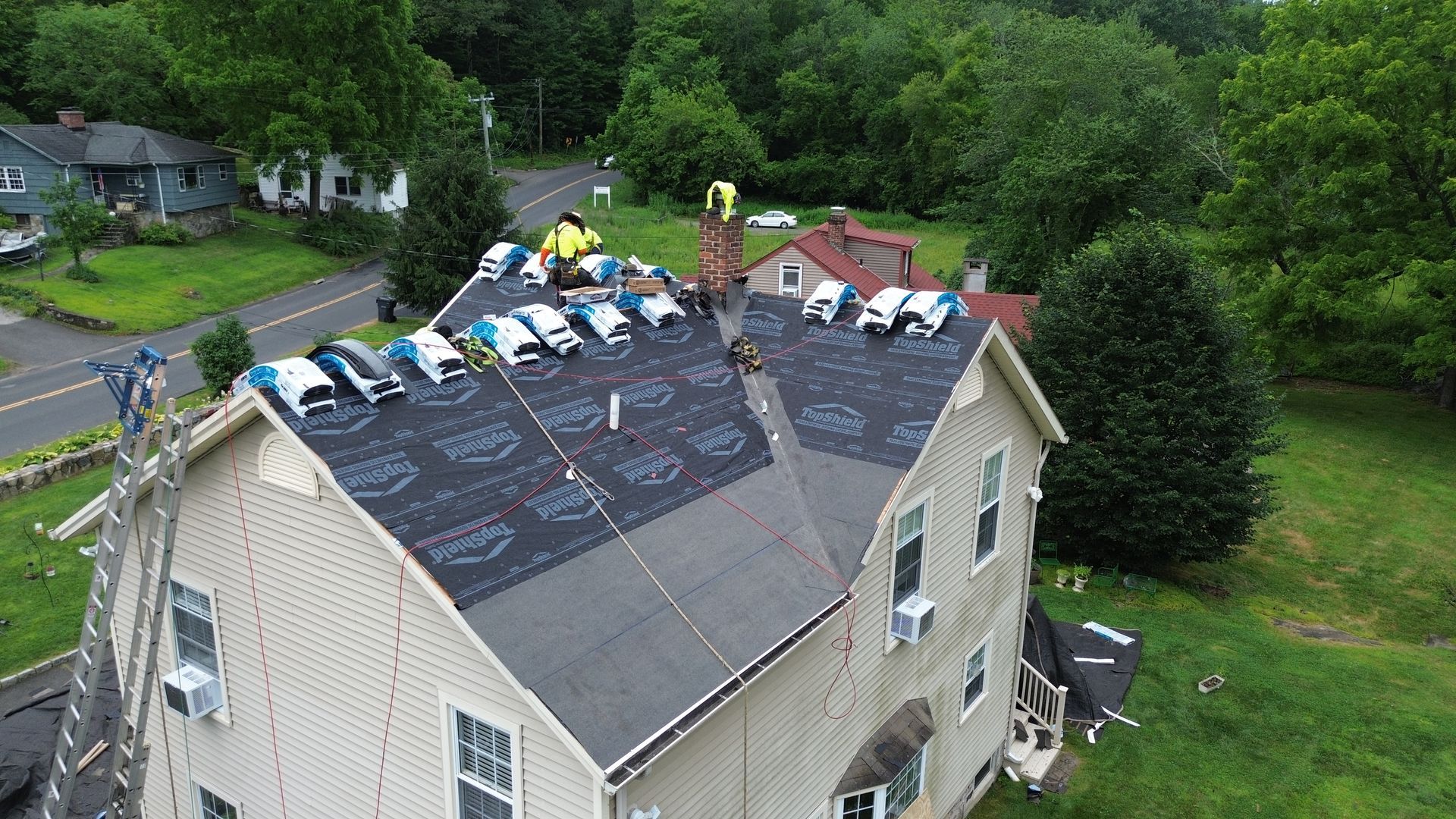 Aerial view of a house with roof damage, covered by tarps, surrounded by trees and grass.