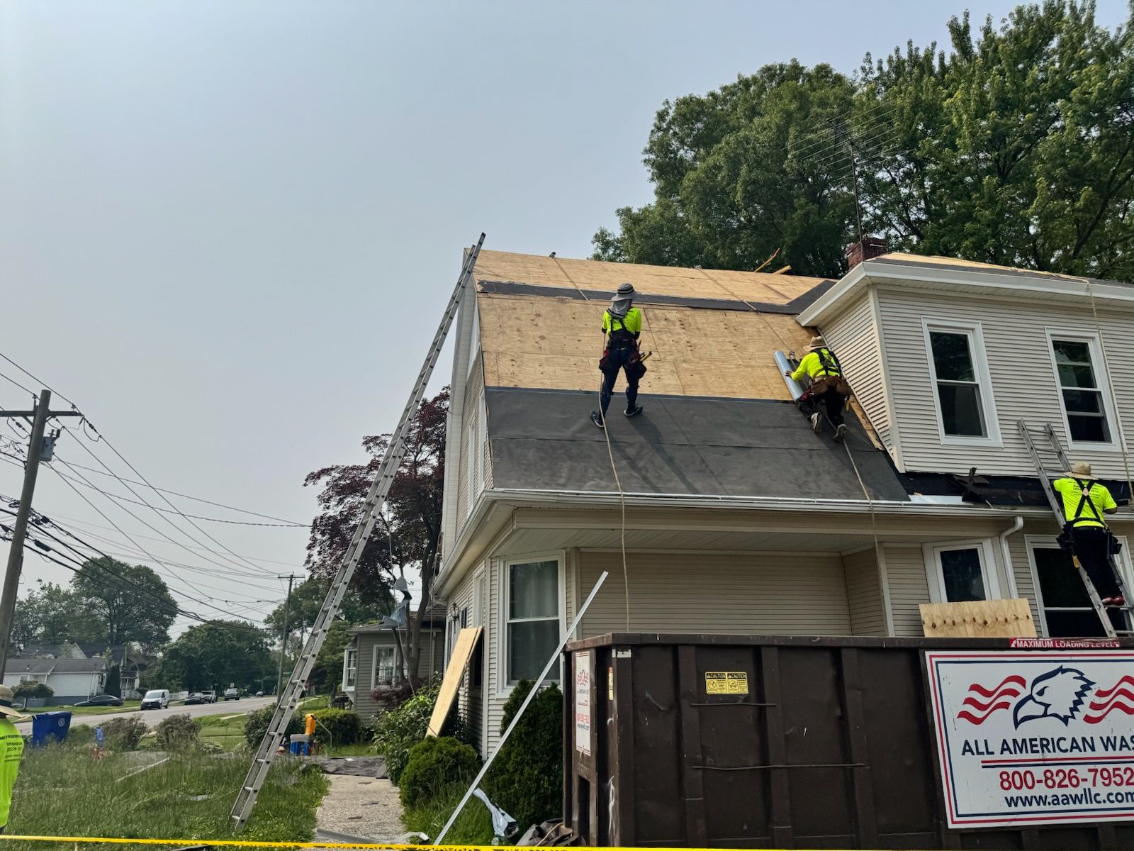 Workers on a house roof replacing shingles beside a tree-lined street.