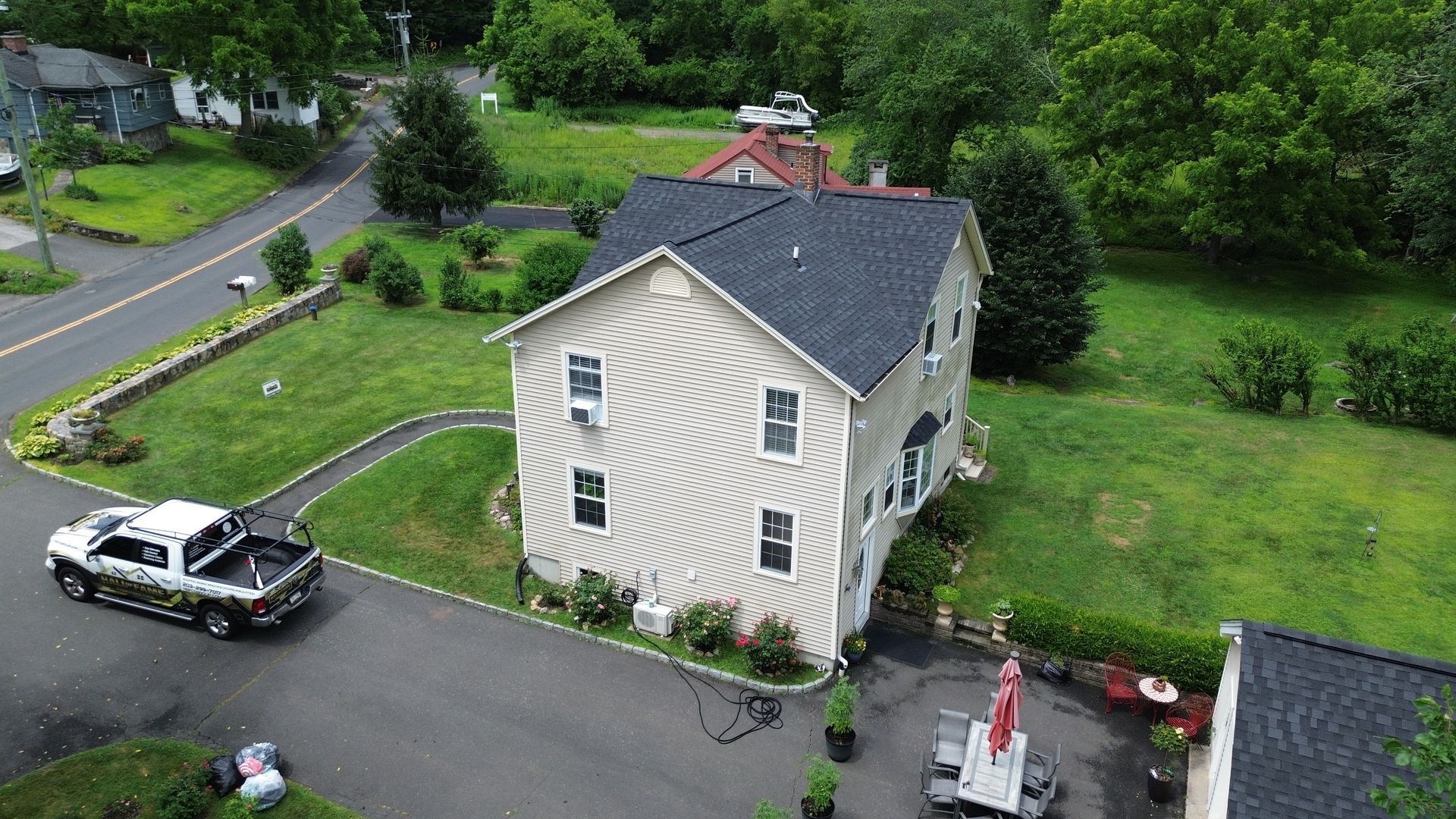 Aerial view of a beige house with a dark roof, driveway, and parked car, surrounded by green lawn and trees