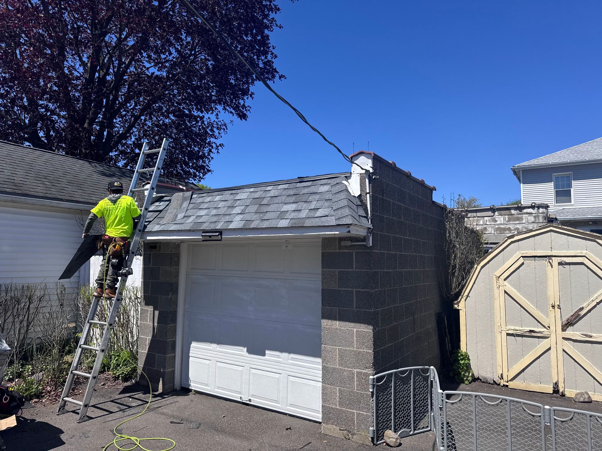 Worker on ladder repairing a small garage roof beside a driveway, with a cinder-block wall and blue sky.