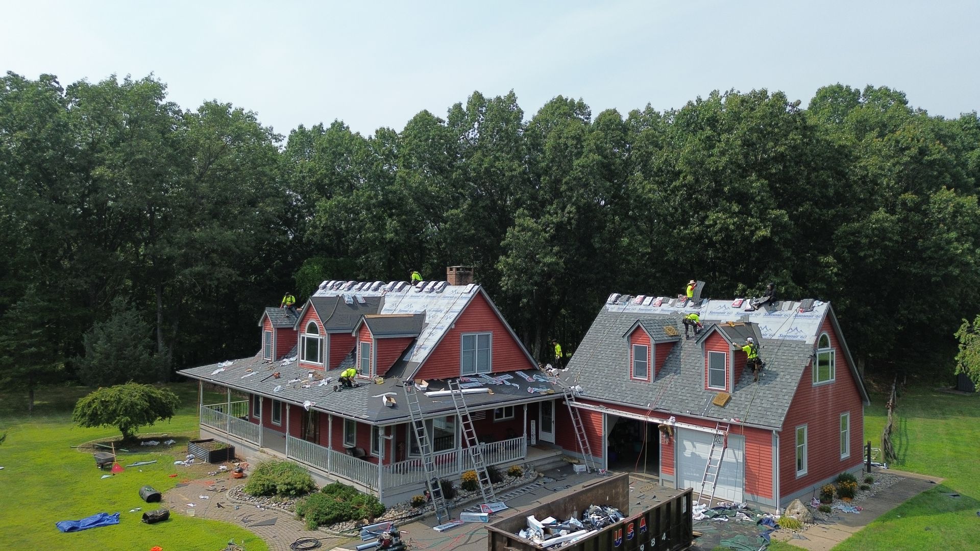Two red houses with gray roofs beside a grassy yard, with people on the porch and trees in the background