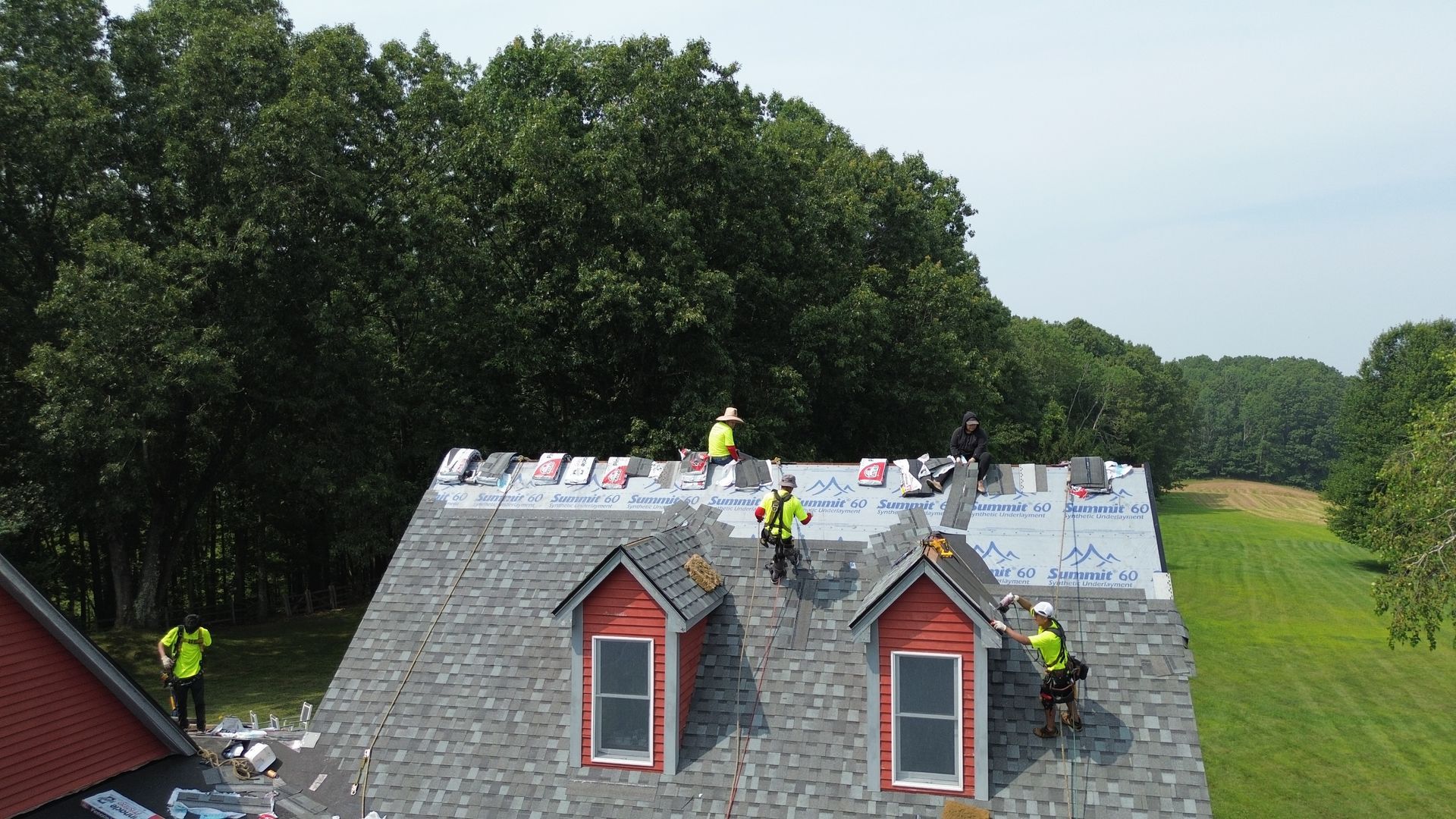 Workers repairing a gray shingled roof with red dormer windows on a house by trees and a field