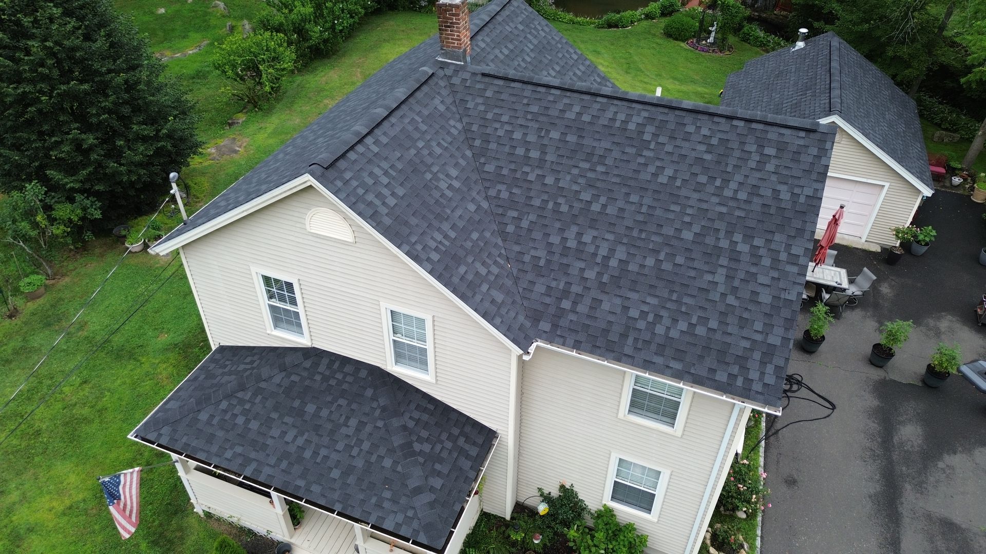 Aerial view of a light beige house with dark gray roof, surrounded by green lawn and a wet driveway.