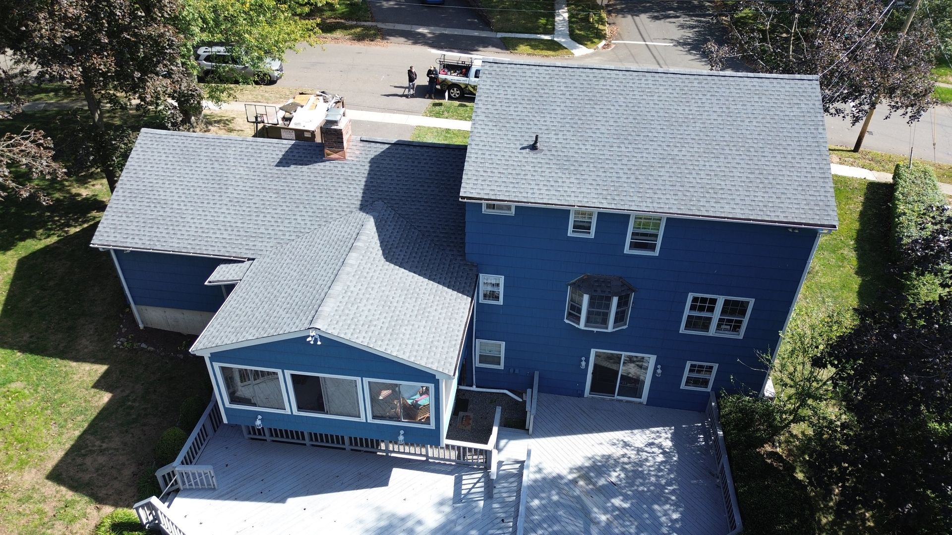 Aerial view of a blue house with gray roofs and a white driveway surrounded by trees and lawn
