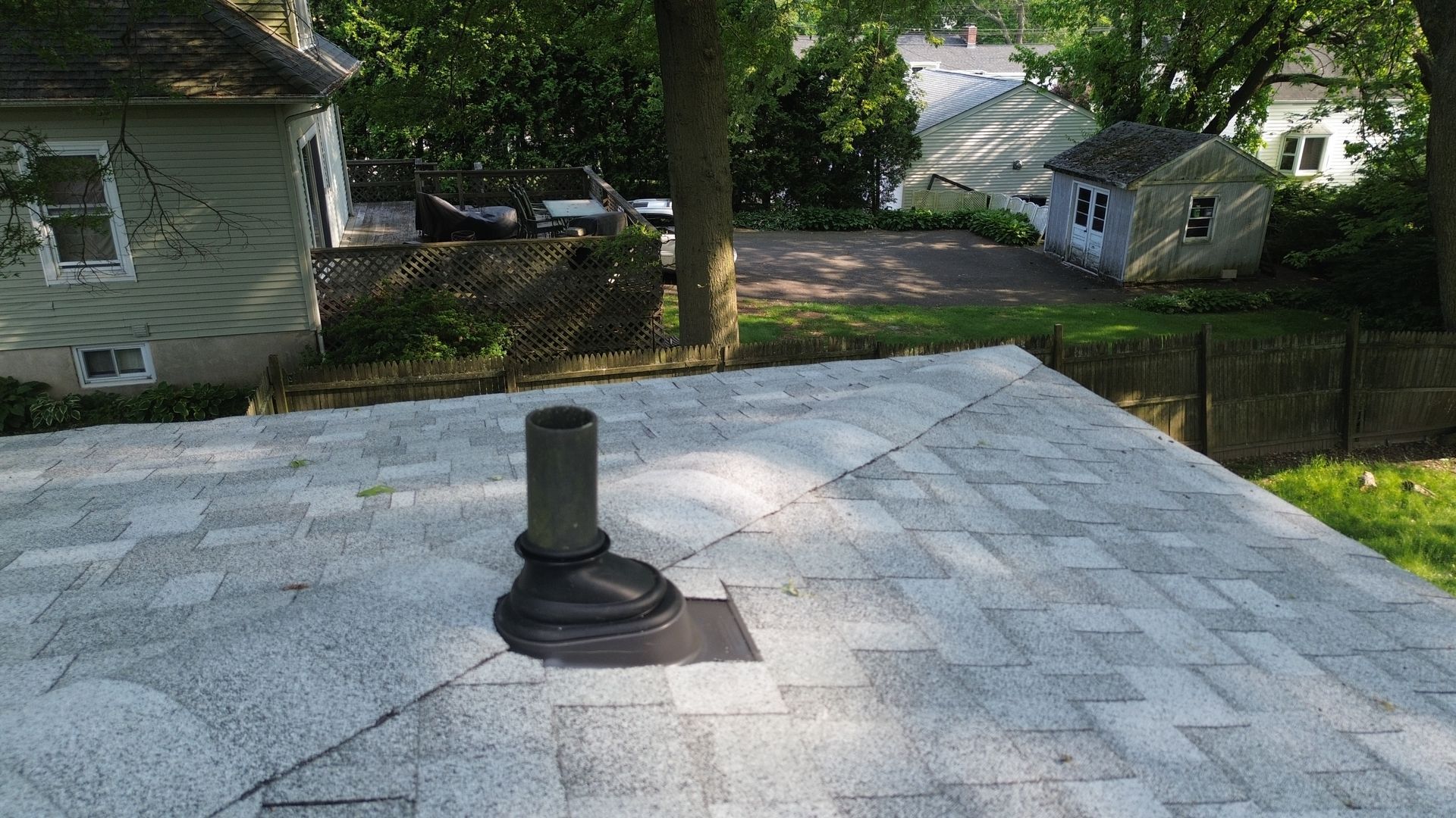 Rooftop with gray shingles and a black vent pipe, overlooking trees and neighboring houses.