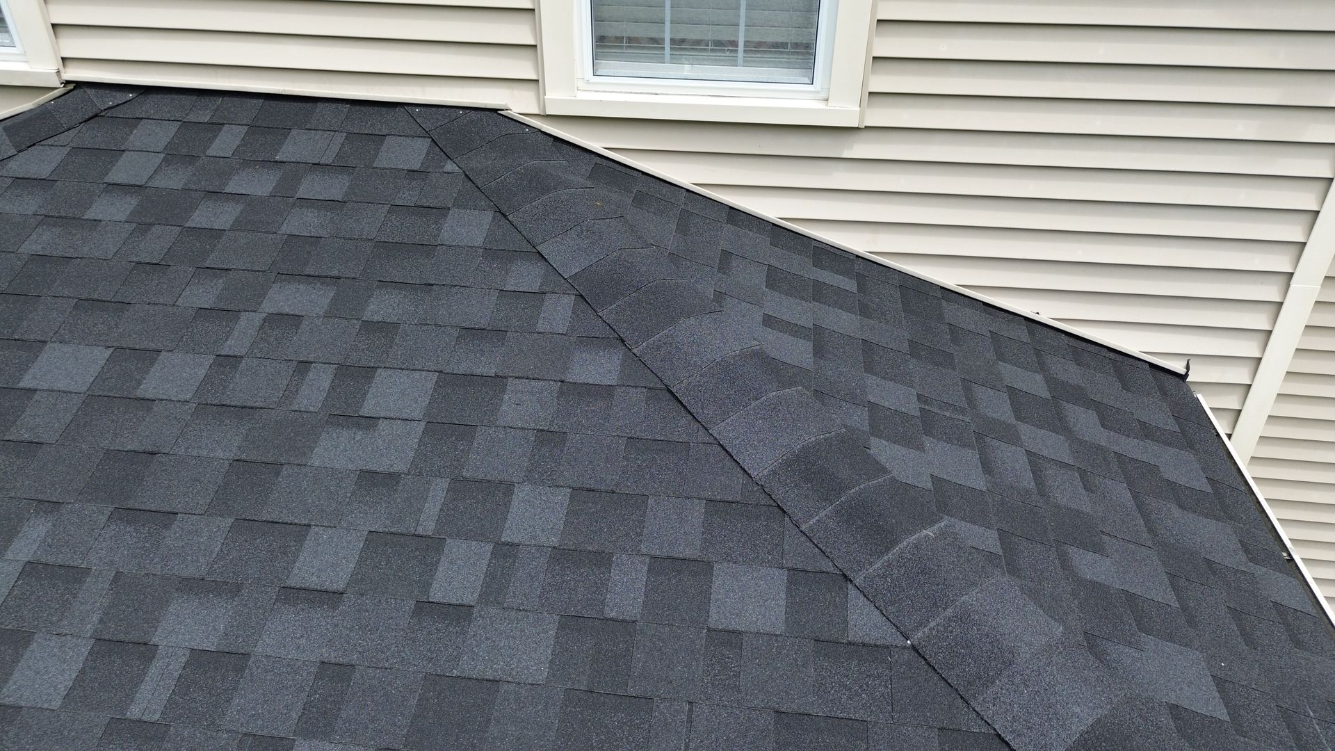 Dark gray asphalt shingle roof on a house, seen close-up against beige siding and windows.