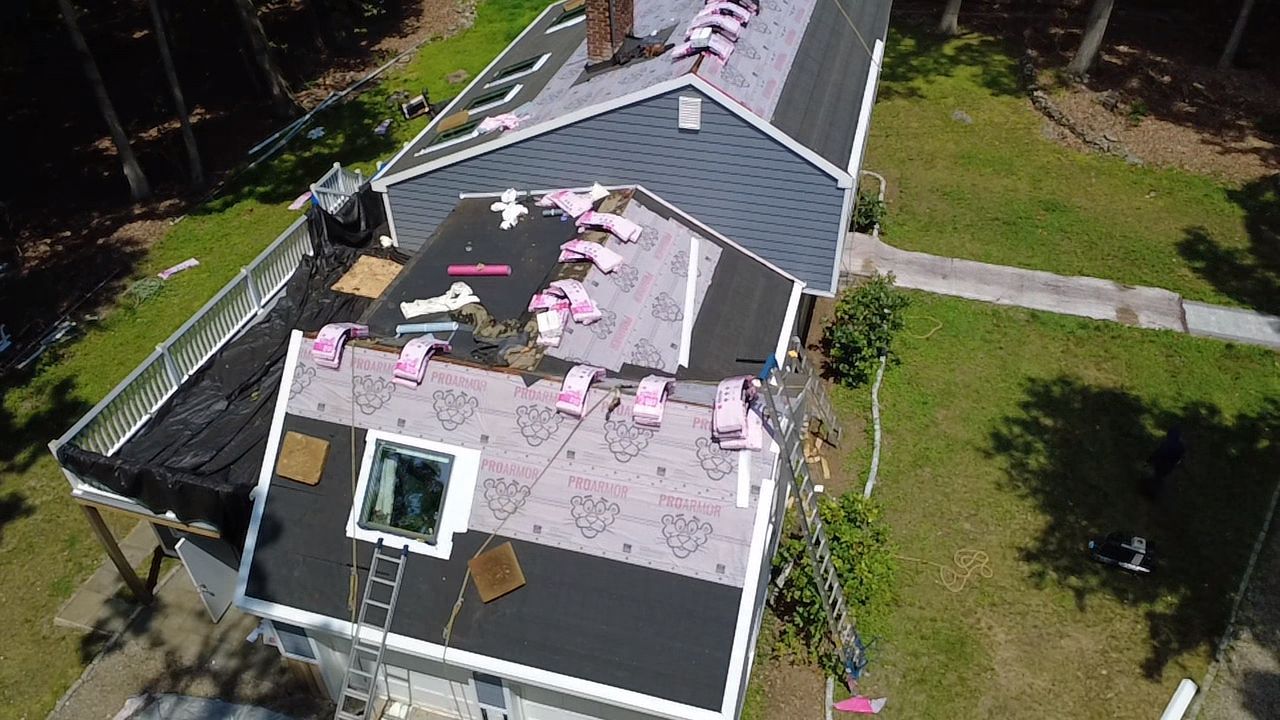 Aerial view of a house with a damaged roof covered in pink and white debris, surrounded by trees and yard.