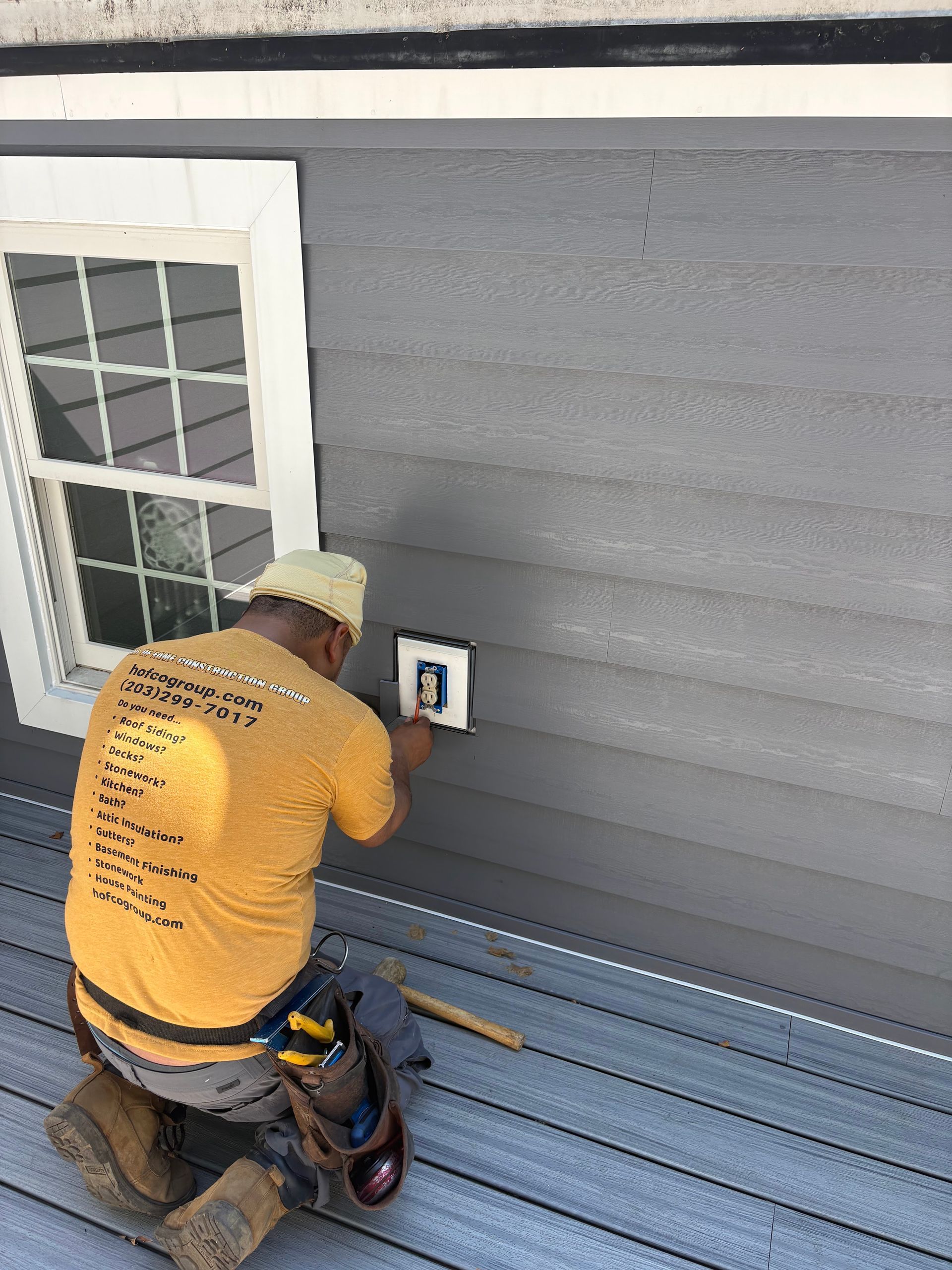 Worker in orange shirt installing an electrical outlet on gray house siding beside a window