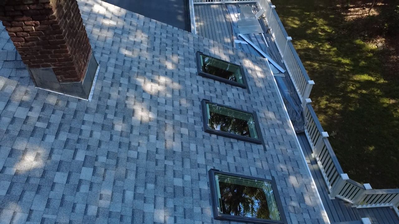 Roof with gray shingles, three skylights, brick chimney, and a white gutter beside trees.