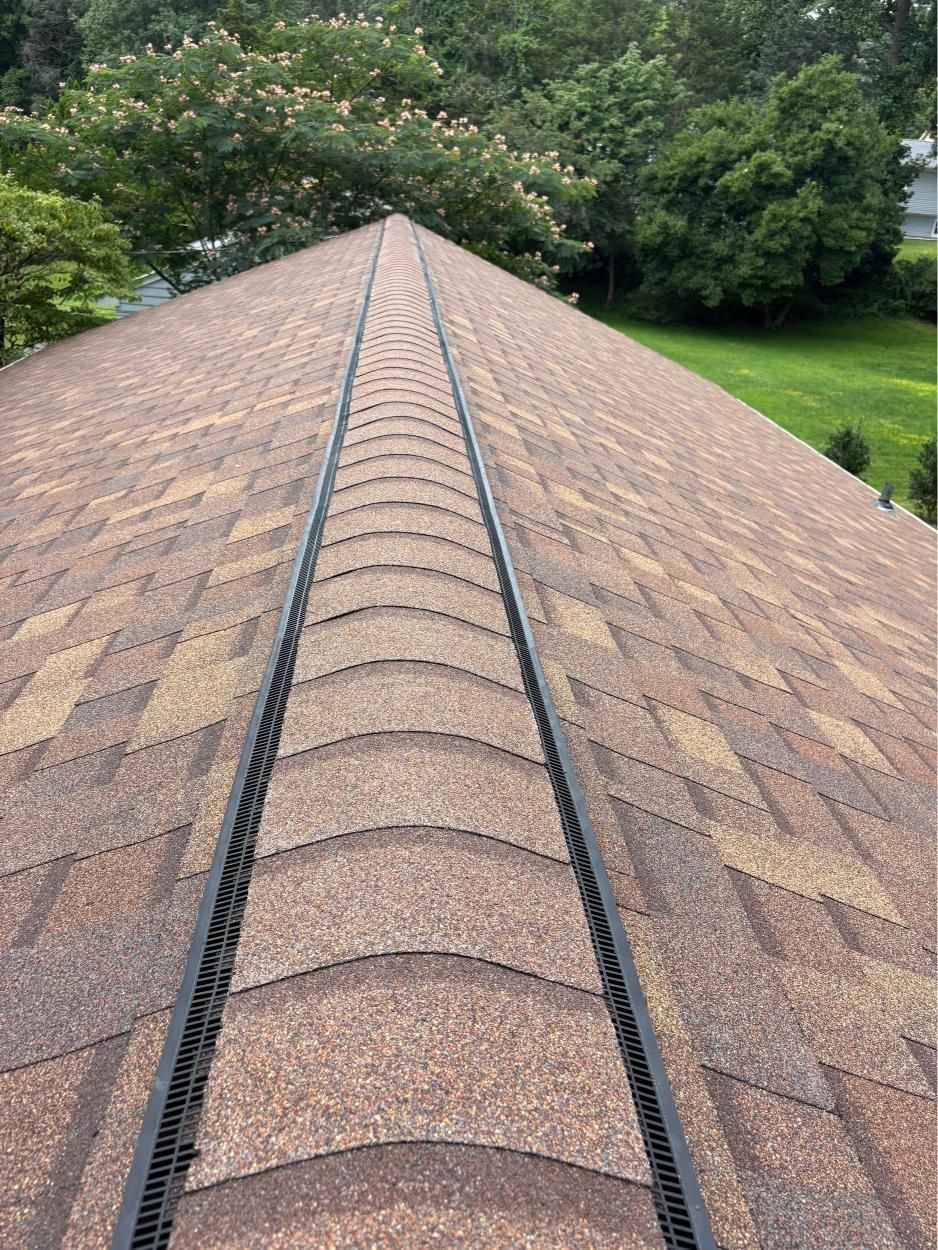 View down a shingled roof ridge with gutters, surrounded by green trees and lawn below.