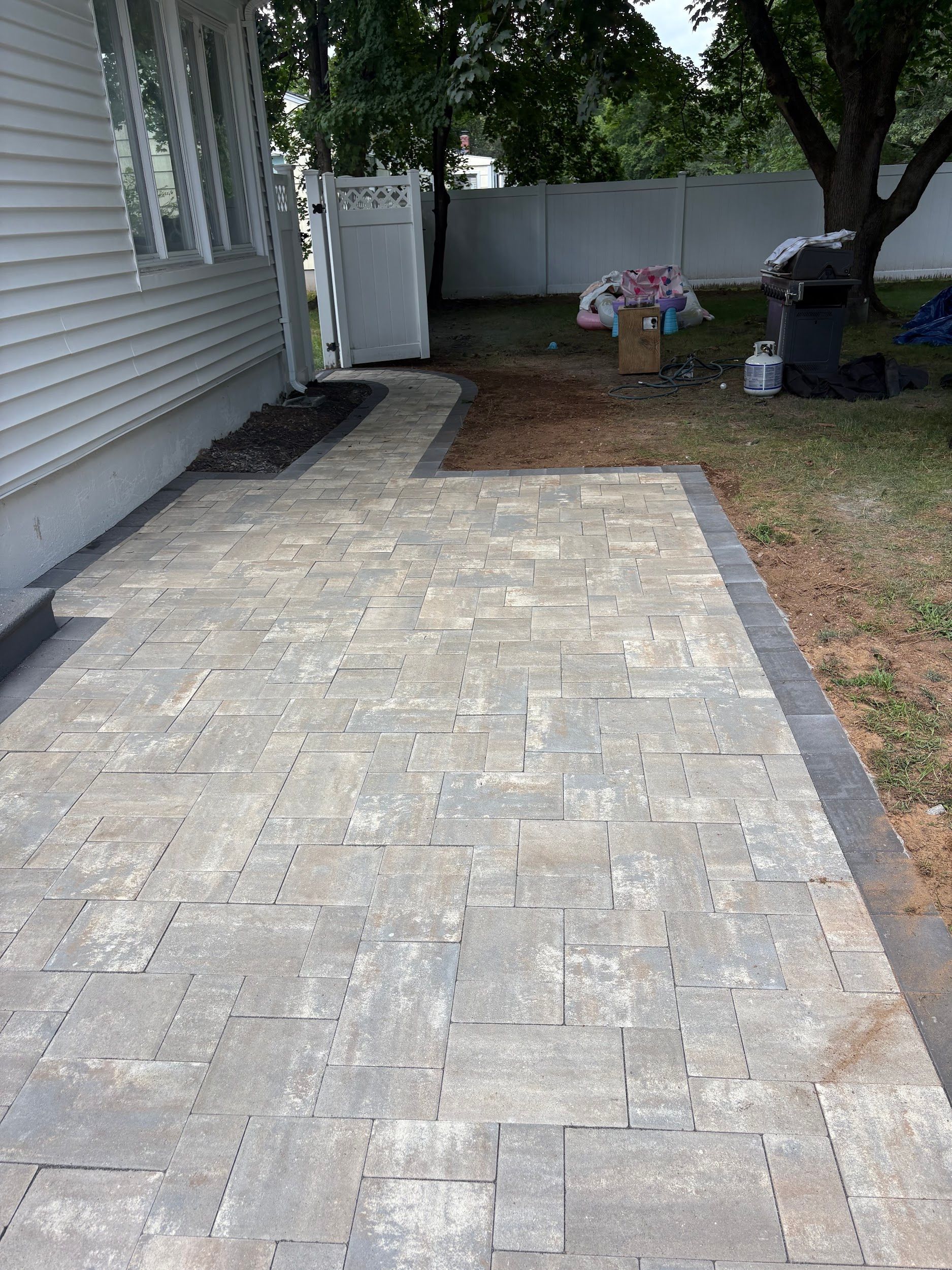 Stone paver walkway beside a house leading to a backyard with trees and lawn