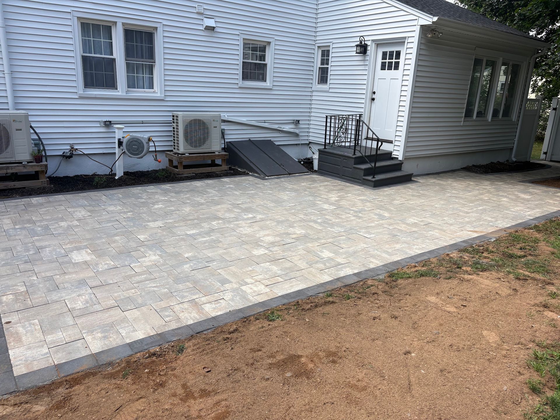 Stone patio beside white house with steps and outdoor HVAC units, bordered by a dirt yard.