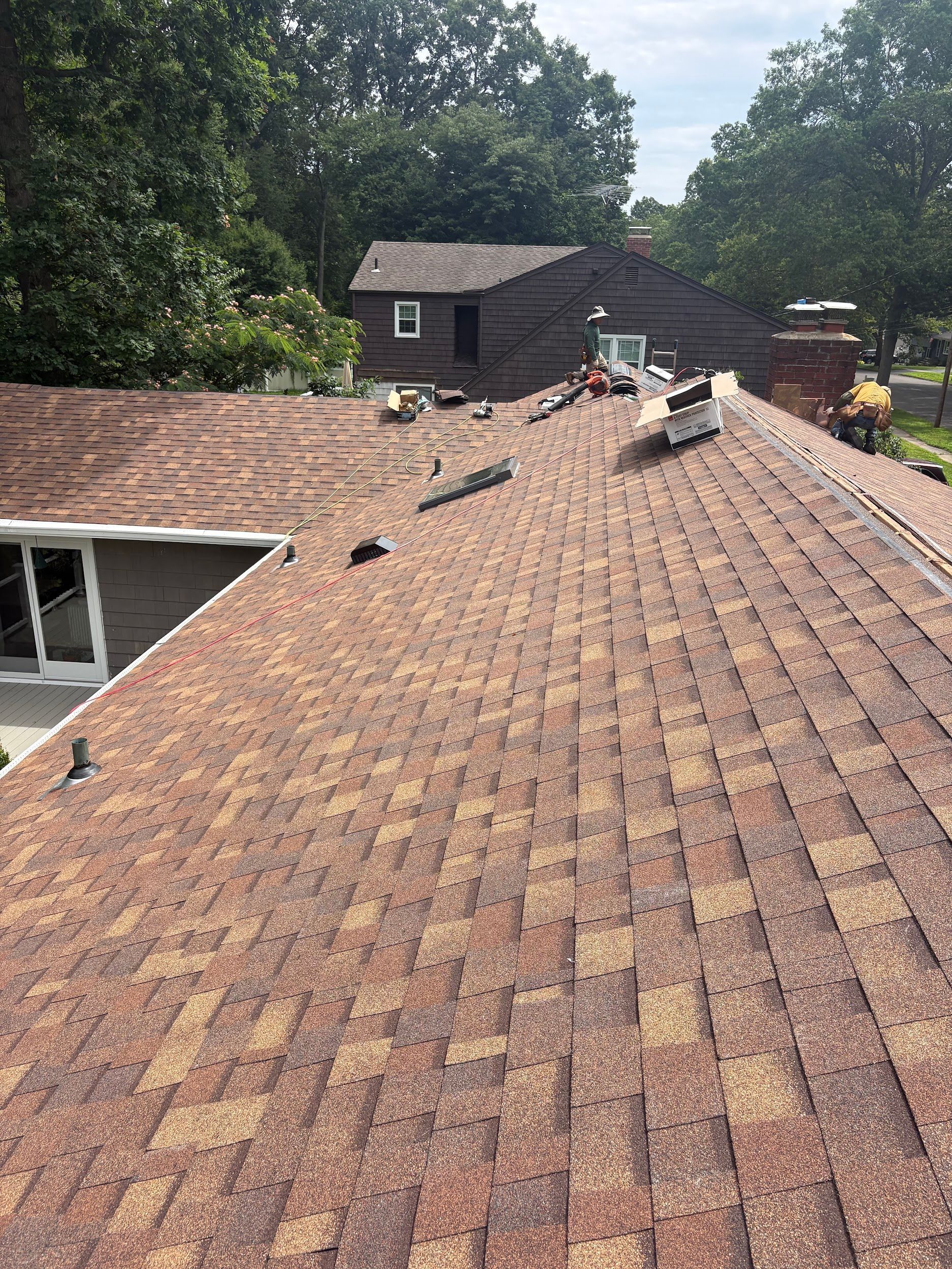 Roofer on a shingle roof with tools and materials, near a house and trees on a sunny day