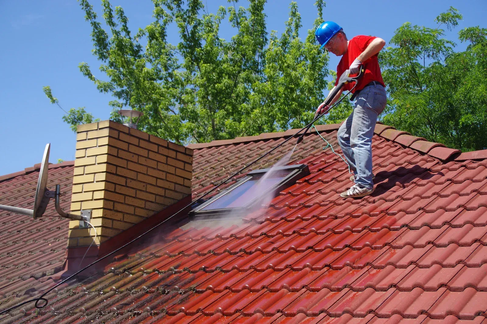 Man on a red-tiled roof using a pressure washer, cleaning. Yellow brick chimney, blue sky, and green trees.