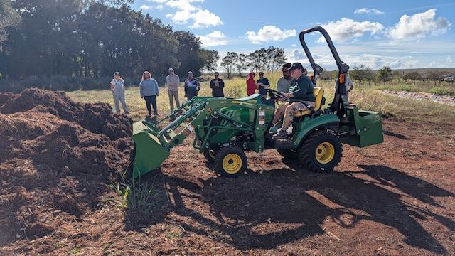 Small tractor with front loader and backhoe working on a dirt field, with people watching nearby