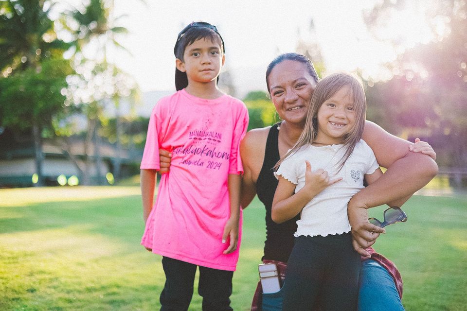 Three people smiling in a sunlit park, with two hugging and one standing nearby in a bright pink shirt