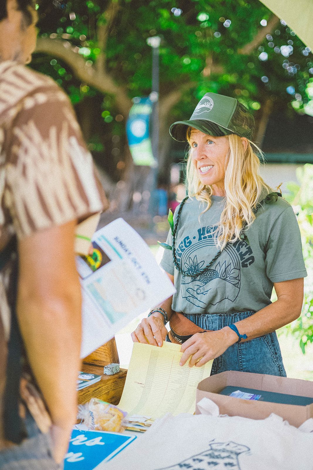 Woman in green cap handing out flyers at an outdoor table under trees