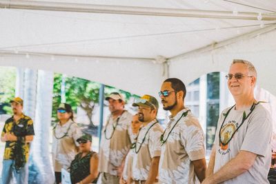 Group of people standing under a white canopy at an outdoor event, wearing matching white shirts and sunglasses