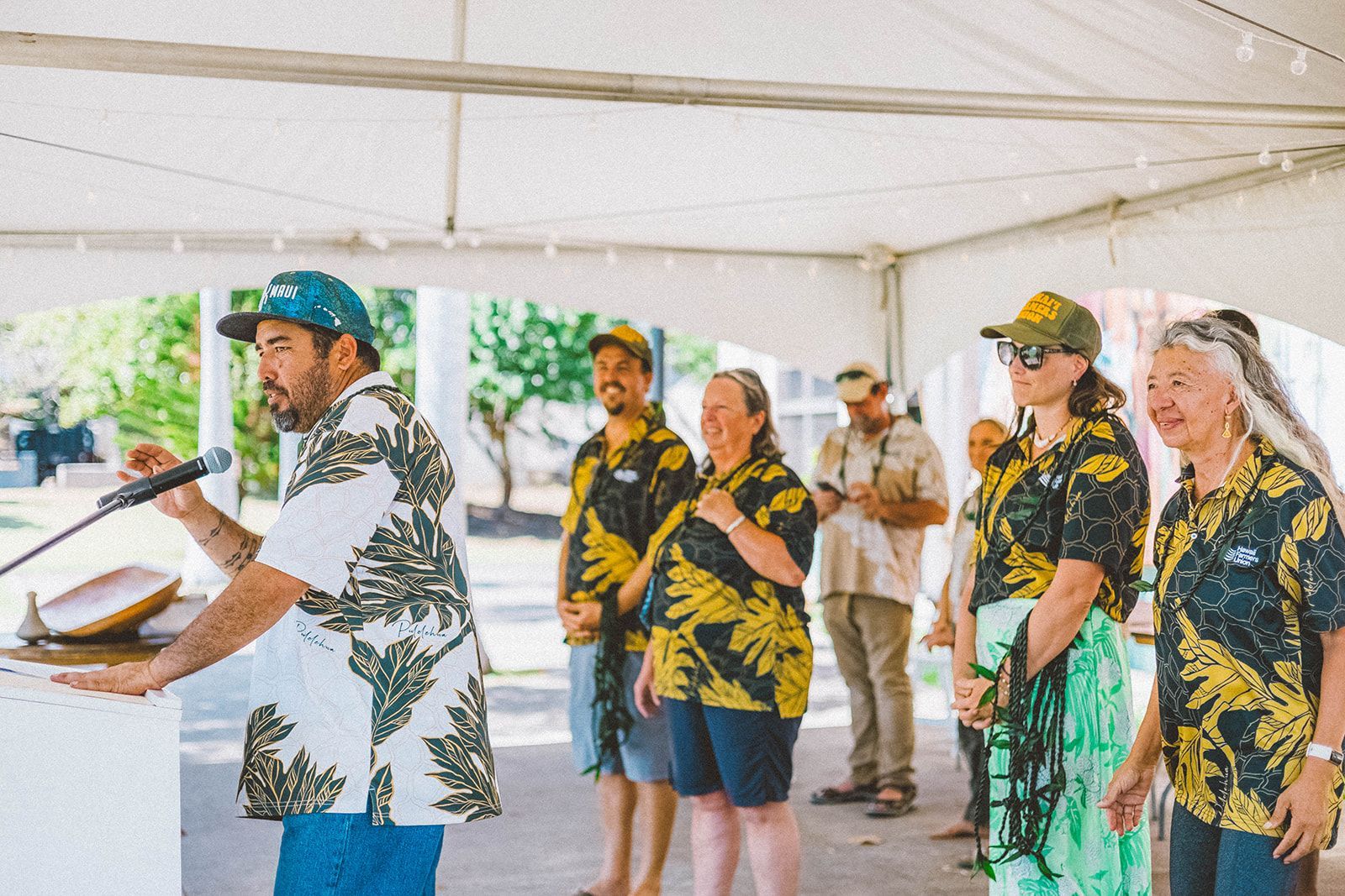 People in matching floral shirts and leis stand under a white tent at a tropical outdoor event