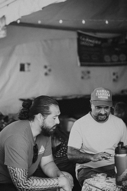 Two men sitting at a table, smiling and talking under a tent at an outdoor gathering.
