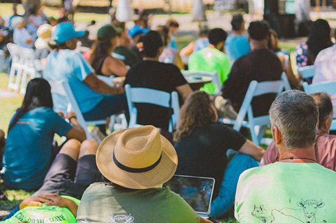 Audience seated outdoors, seen from behind, with a straw hat in the foreground.