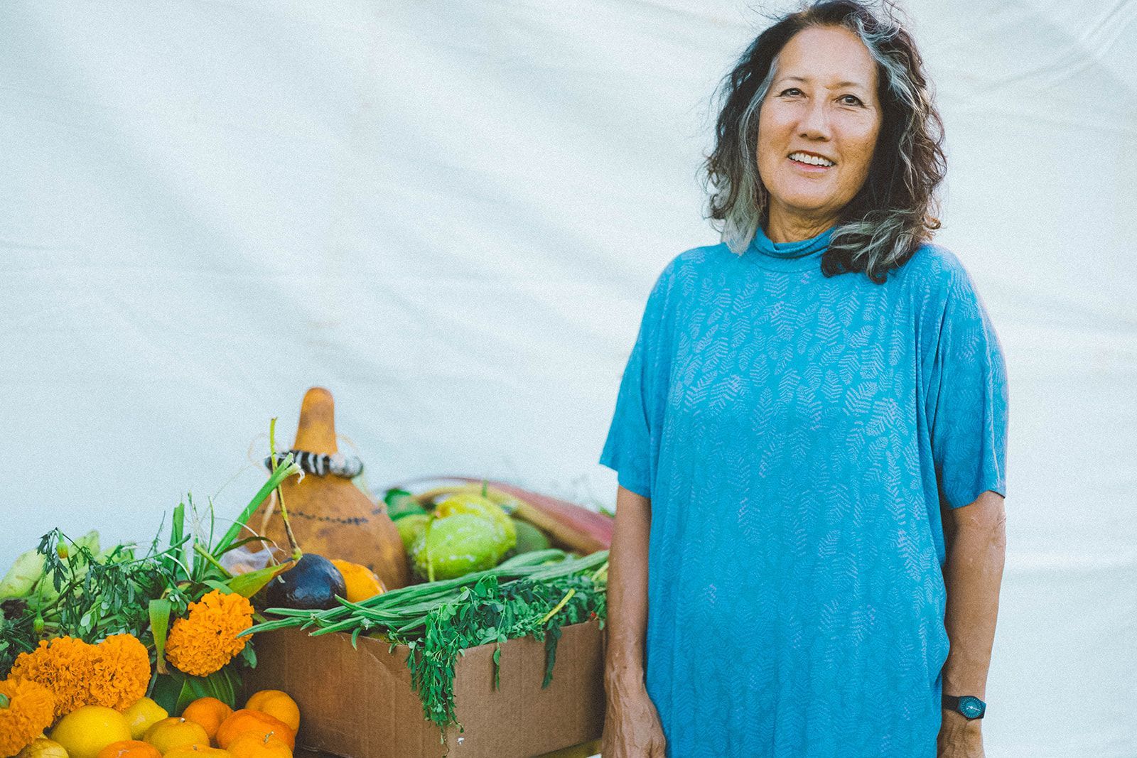 Smiling person in a blue dress stands beside a basket of fruits and flowers