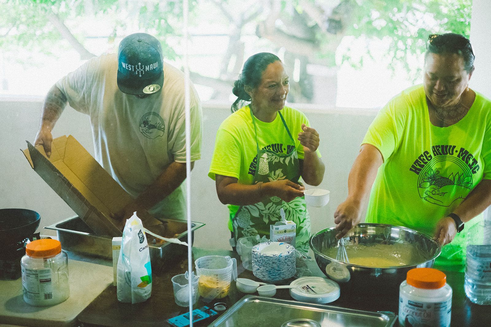 Three people cooking at an outdoor table, wearing bright green shirts beside pots and ingredients.