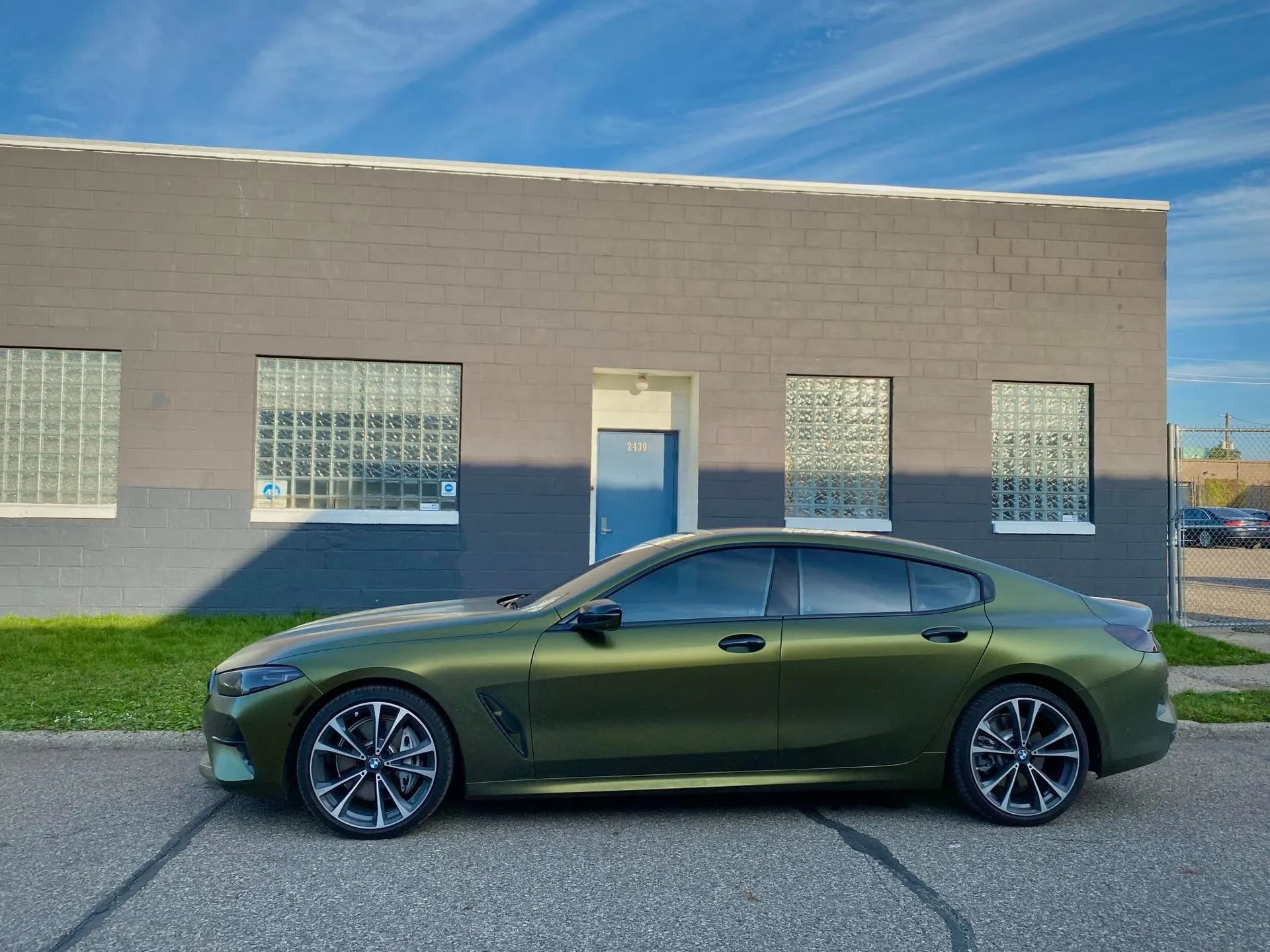 Olive-green BMW sedan parked in front of a gray building on a sunny day.