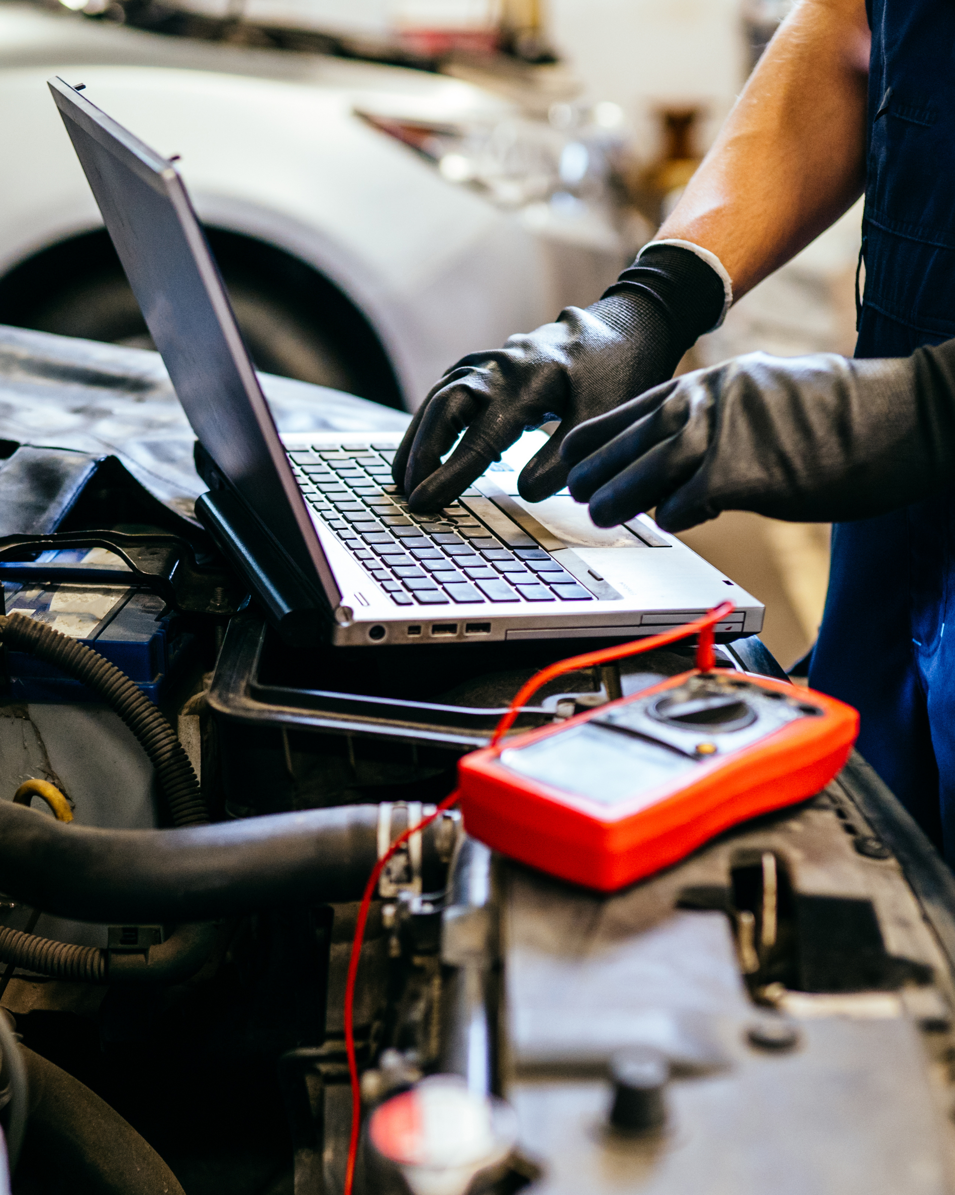 Mechanic in gloves using laptop and multimeter on a car engine.