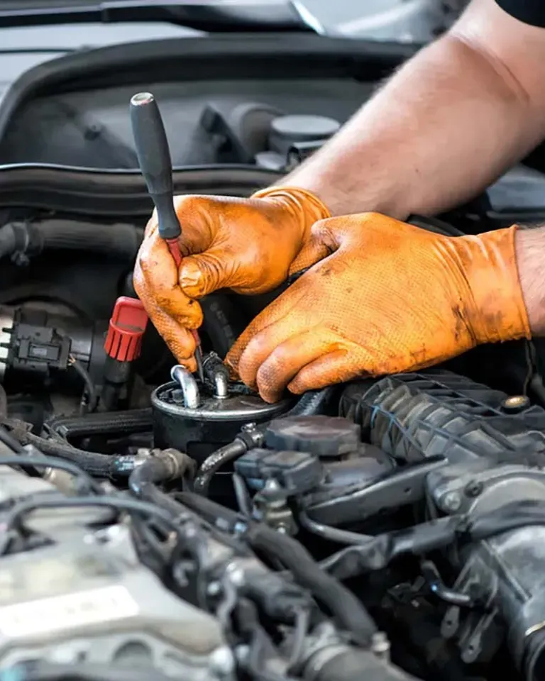 Hands wearing orange gloves working on a car engine with a screwdriver.